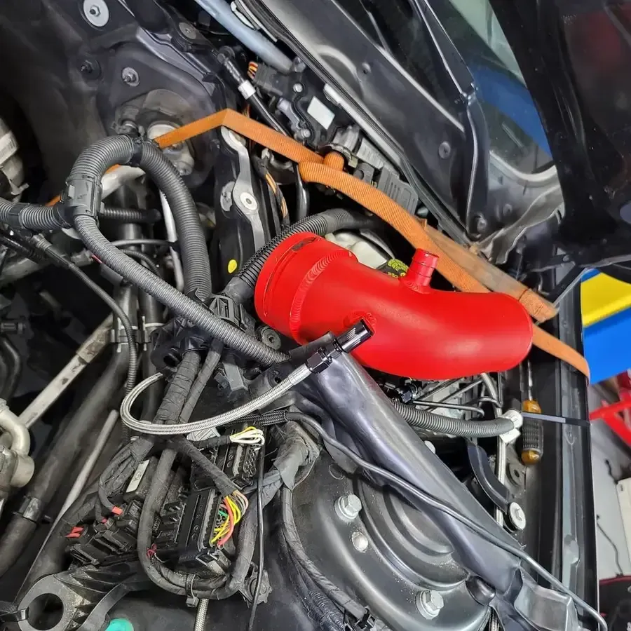 Red automotive intake tube installed in a car engine bay, with wiring and other parts visible.