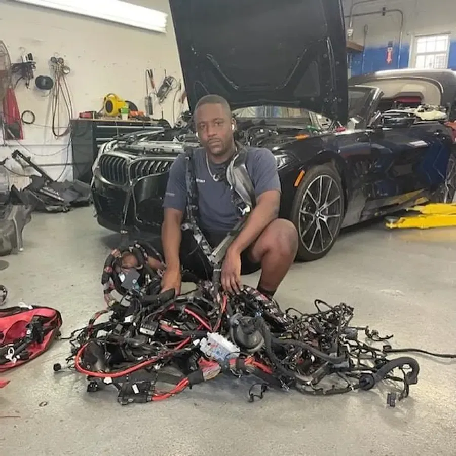 Man kneeling in a garage with car and wires. Black car with hood open behind him. Wires are in a pile.
