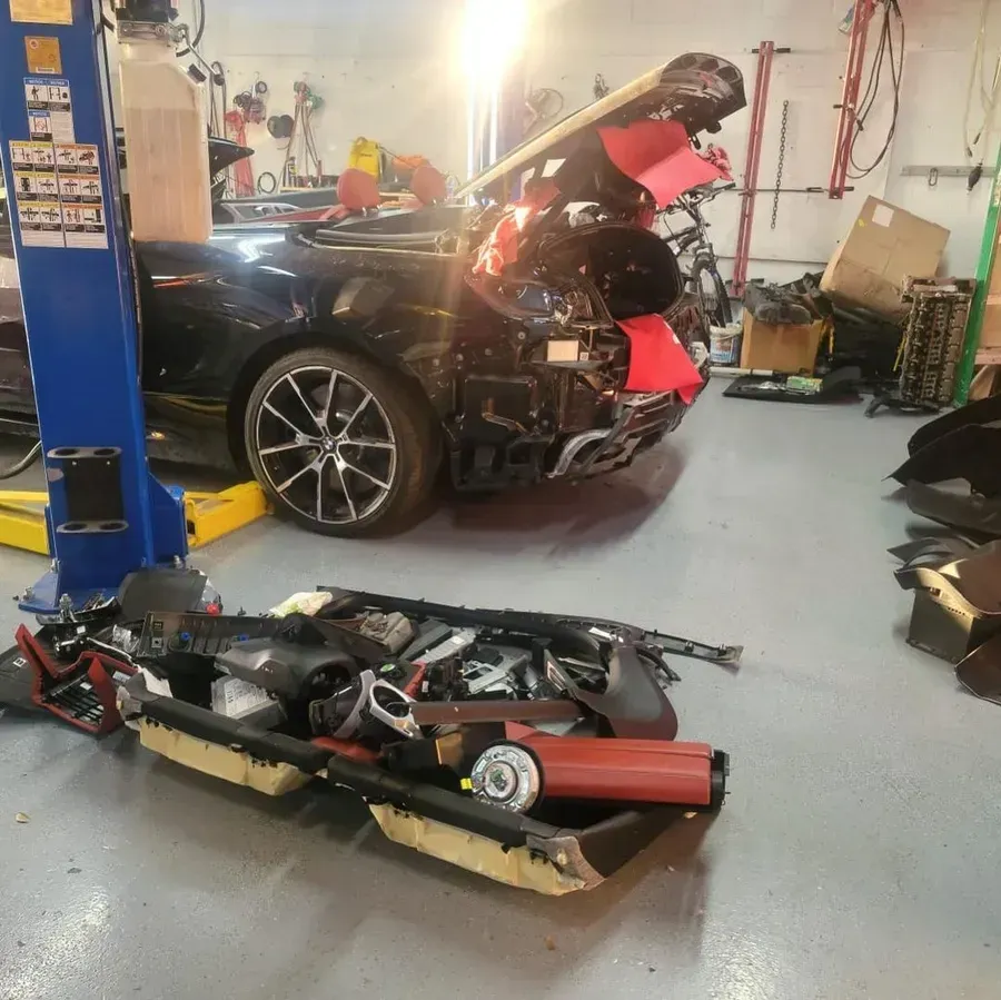 Damaged black car in a repair shop with parts scattered on the floor and body panels removed.