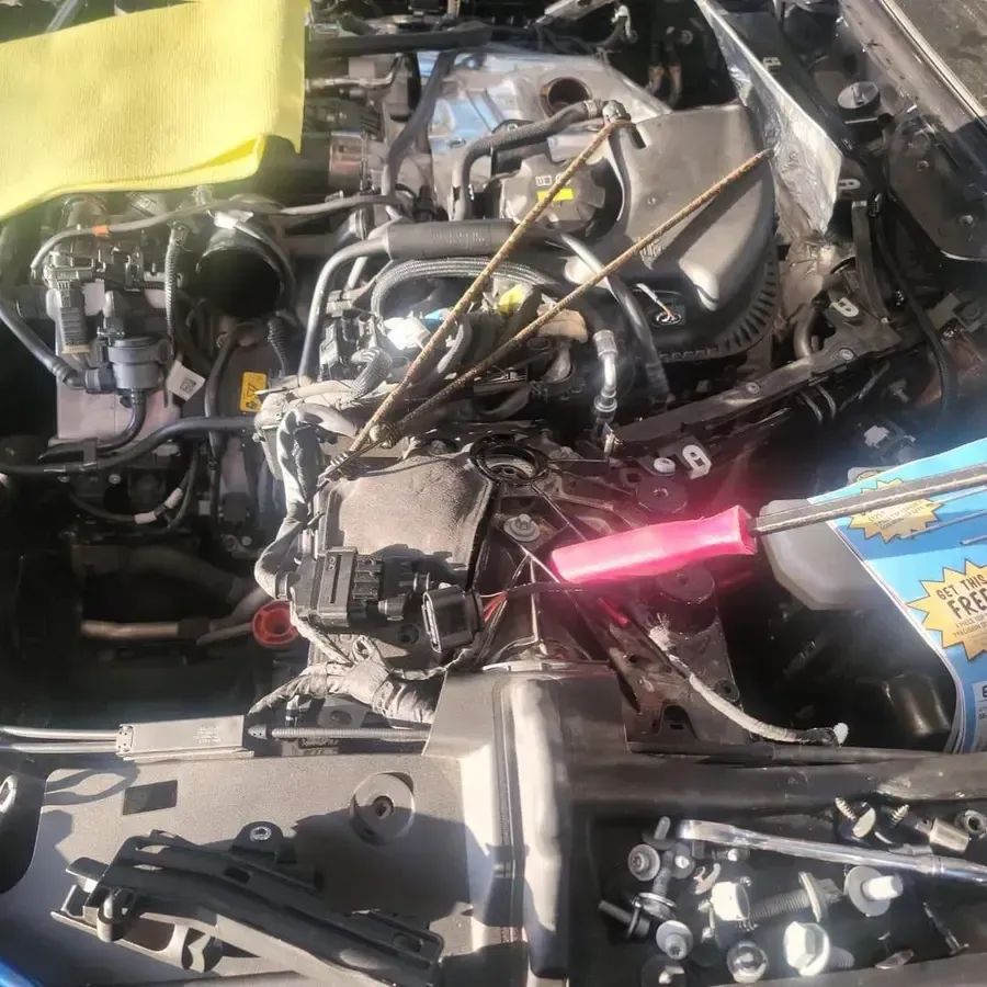Car engine bay with parts removed, tools, and a yellow cloth, lit by sunlight.