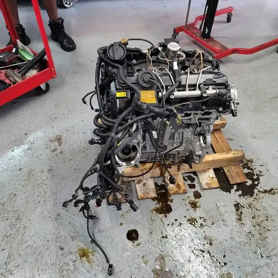 Car engine on a wooden pallet in a garage, with wiring and fluids visible.