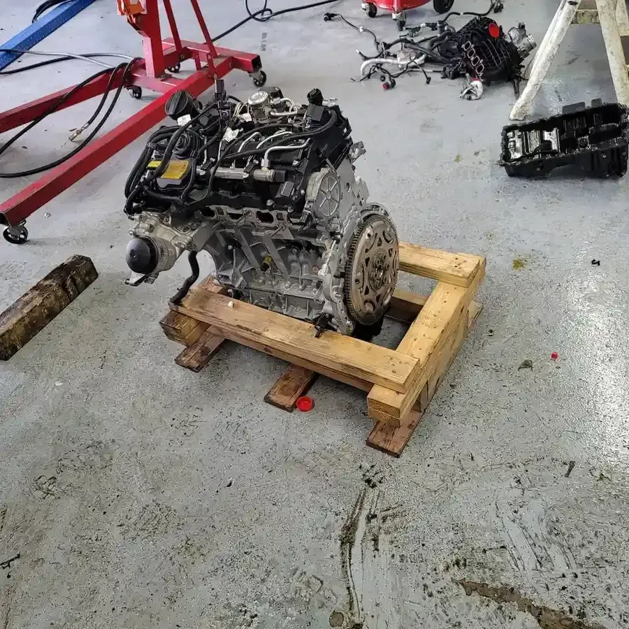 Engine on a wooden pallet in a garage, surrounded by automotive parts and tools.