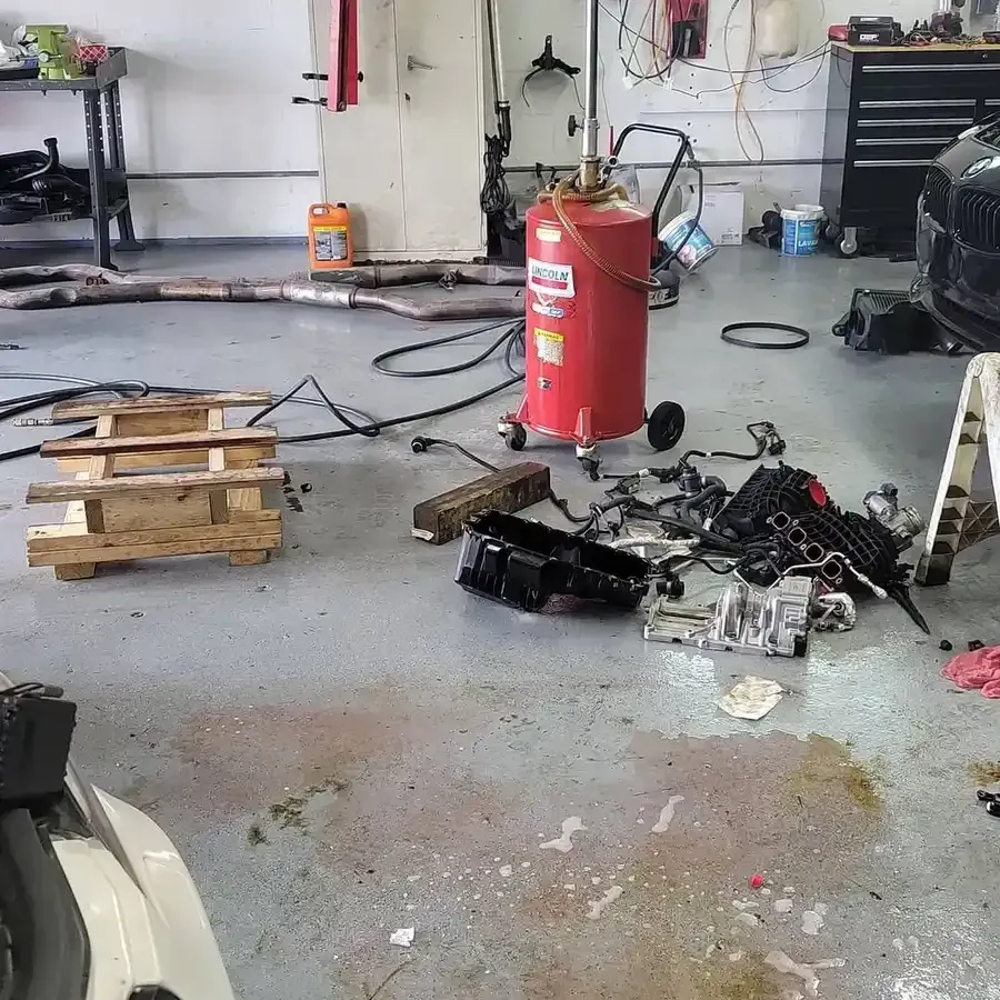 Mechanic shop with scattered car parts, oil stains on the floor, and equipment like a red fluid tank and a toolbox.