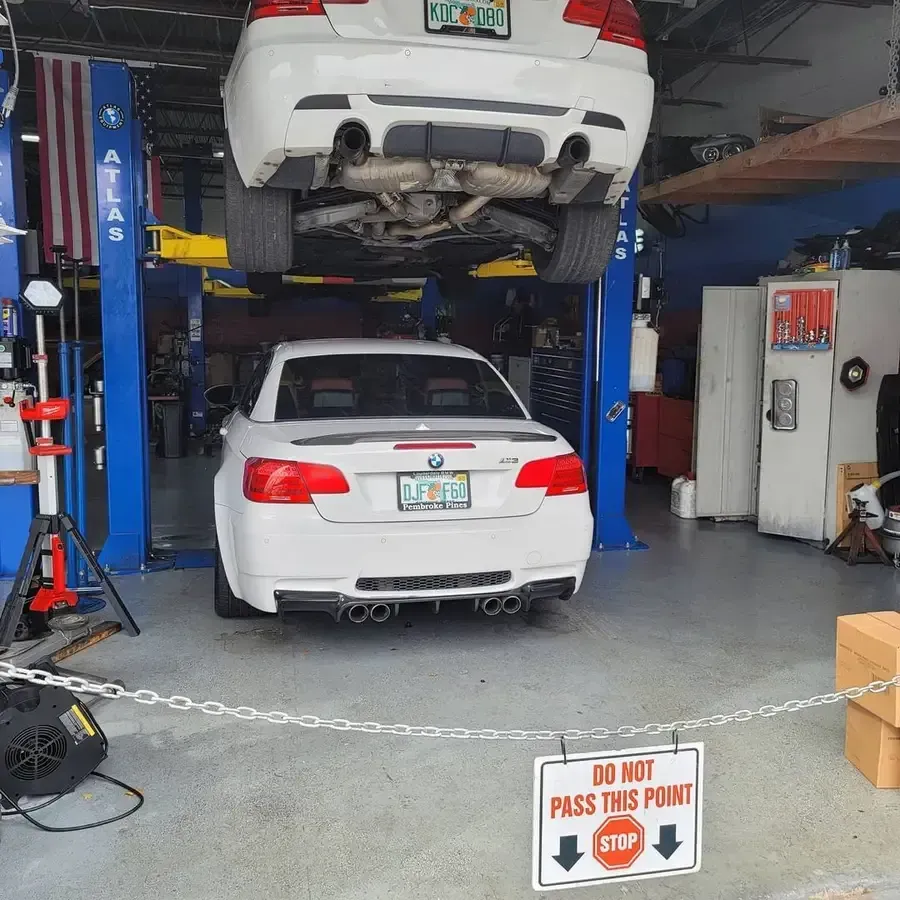 Two white BMWs in a repair shop, one on a lift, the other on the ground. A "Do Not Pass" sign is in front.