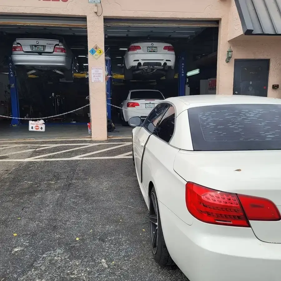 White BMW convertible parked outside a car repair shop with other BMWs on lifts.