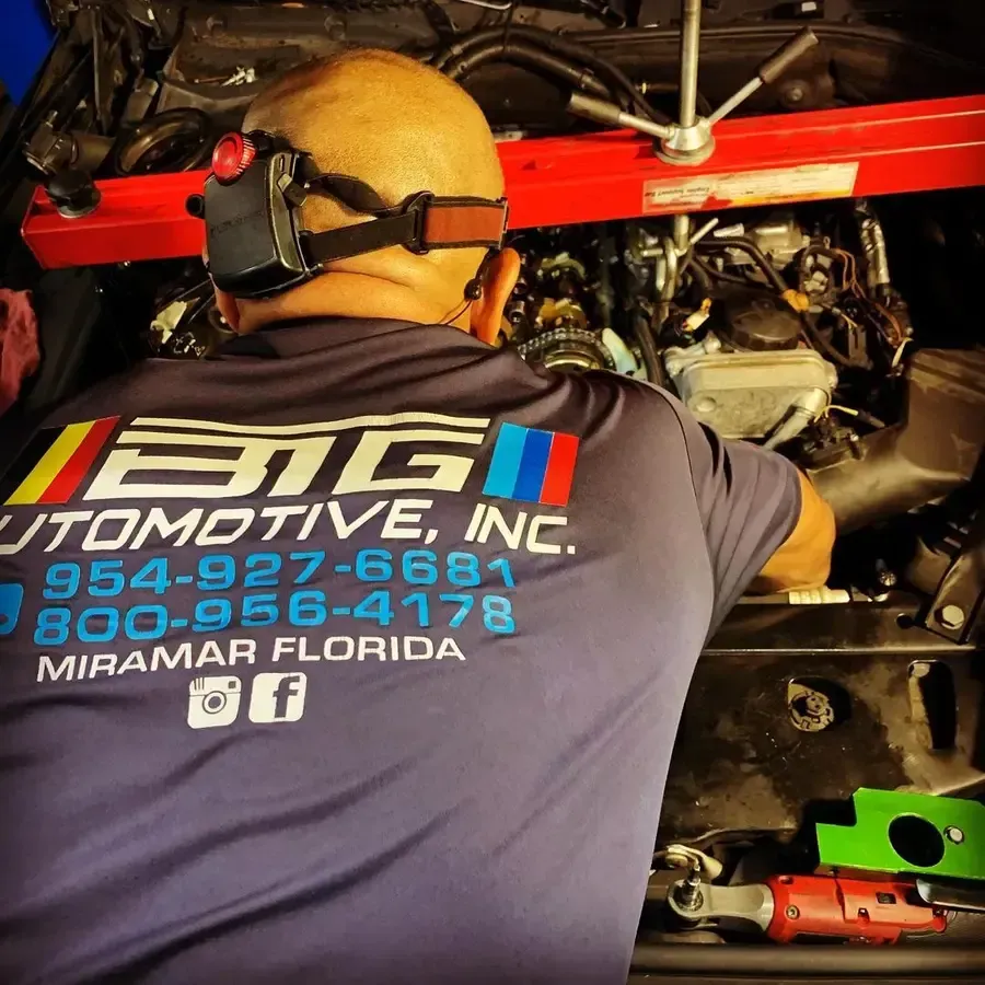 Mechanic working on car engine, wearing earmuffs. BMG Automotive, Inc. logo on shirt. Miramar, Florida.