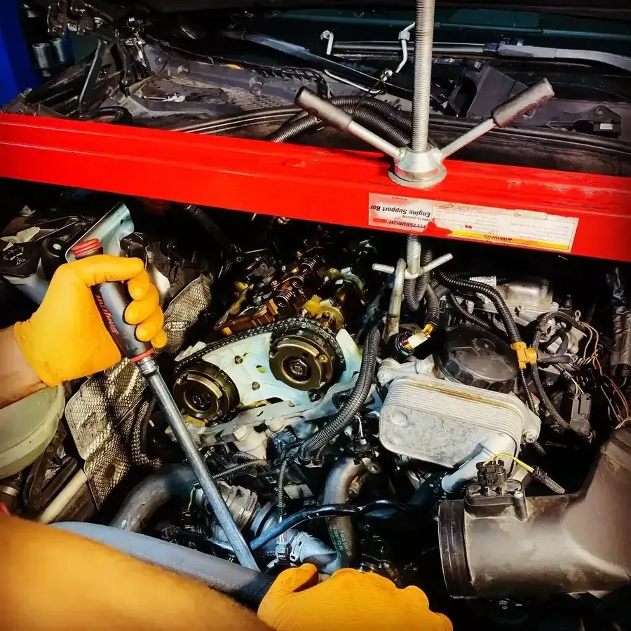 Mechanic working on car engine, using tool. Orange gloves and red support beam visible.