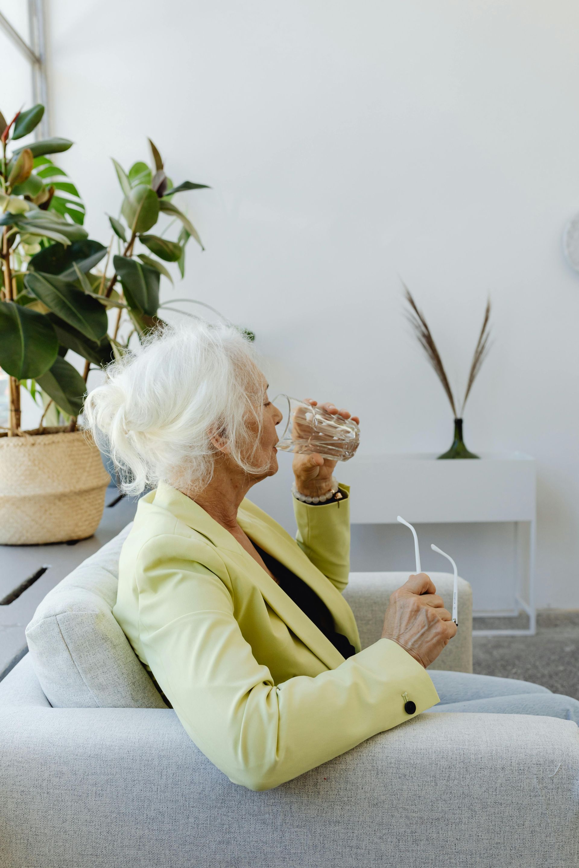 An elderly woman is sitting on a couch drinking a glass of water.