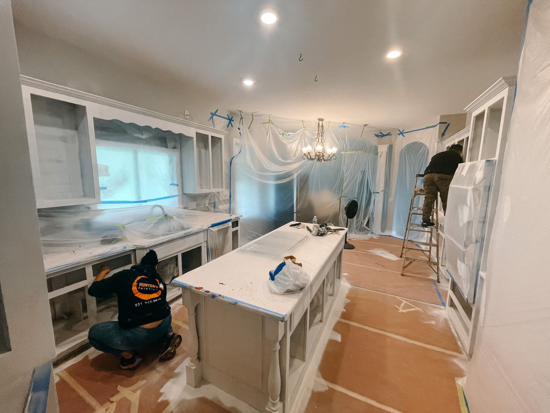 A man is kneeling down in a kitchen painting cabinets.