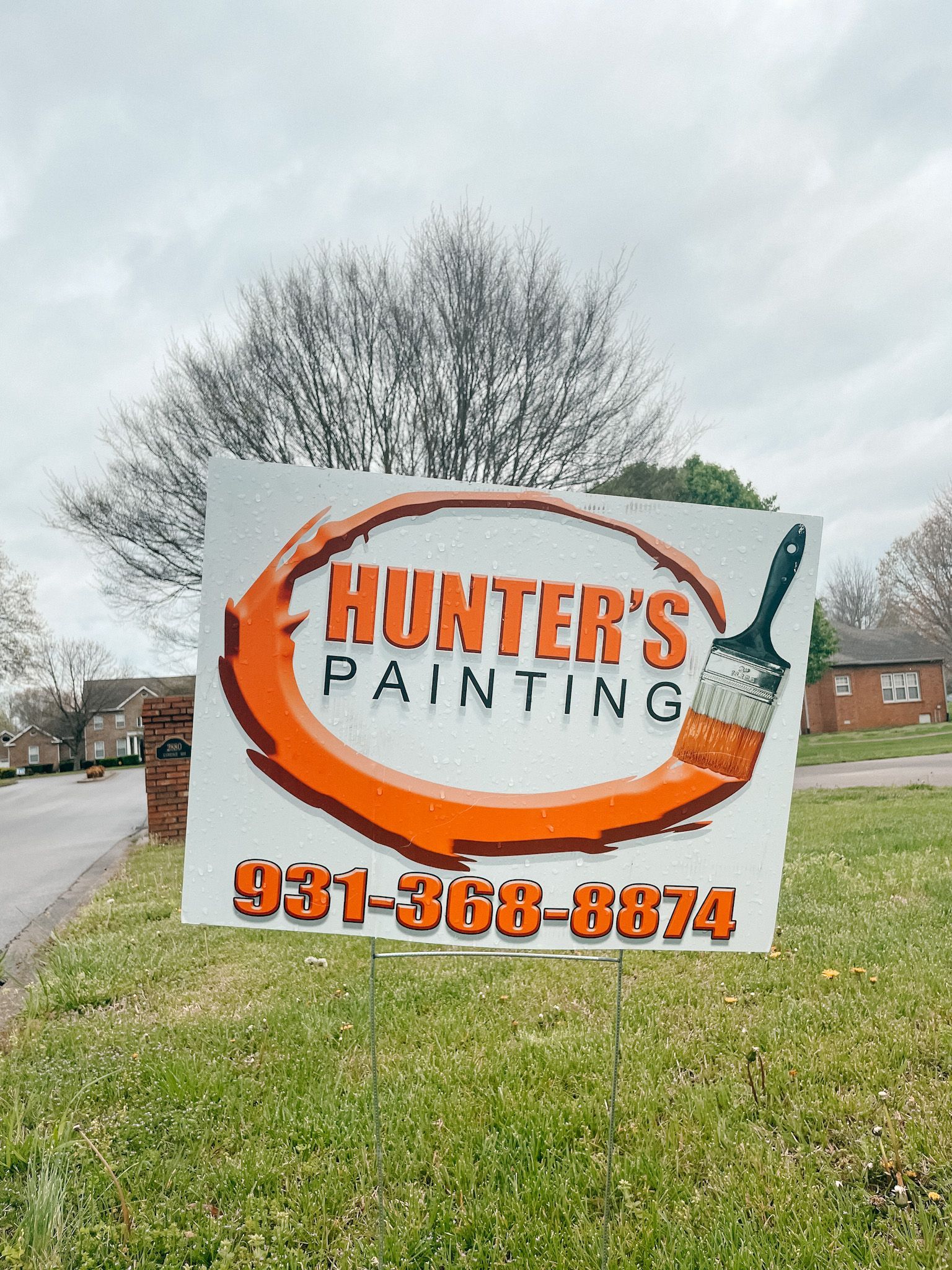A sign for hunter 's painting is sitting in the middle of a grassy field.