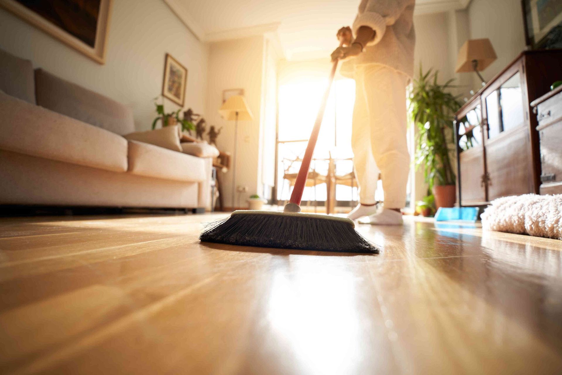 A person in white clothing uses a broom to clean a hardwood floor in a sunlit living room.