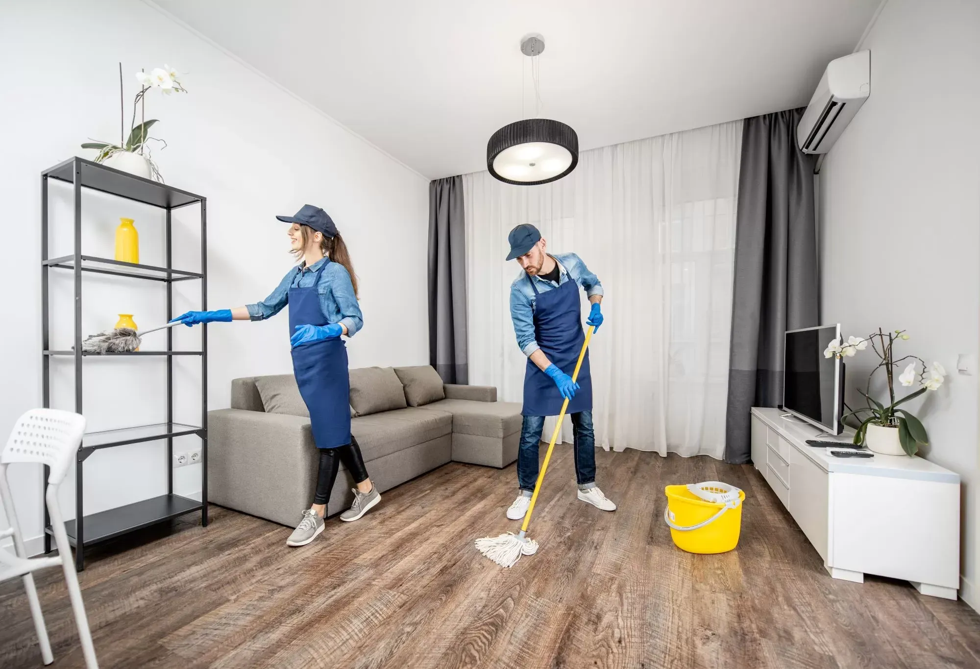 Two people in blue uniforms and gloves clean a living room: one wipes a tall metal shelf while the other mops the floor.