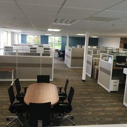 An office interior featuring a central conference table with six chairs, surrounded by rows of grey office cubicles.