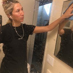 A person wearing a black shirt and headphones wipes a wet, spotted bathroom mirror with a blue cloth.