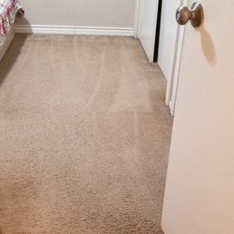 A view of a carpeted room from a doorway, showing a hallway leading toward a partially visible bed on the left.