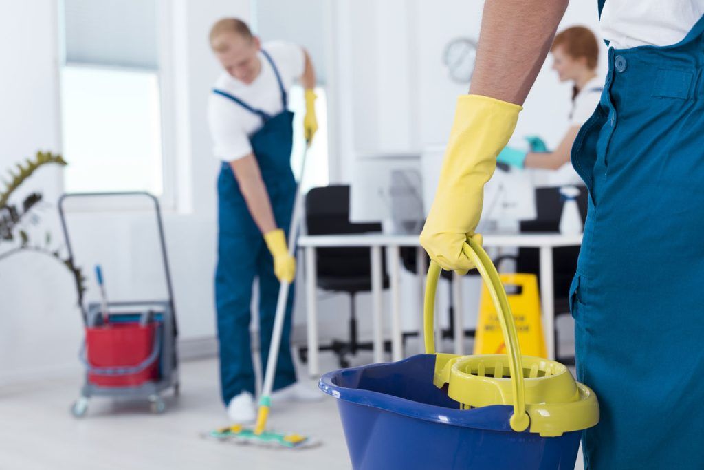 A team of cleaners in blue jumpsuits and yellow gloves cleans an office, with one foreground worker holding a mop bucket.