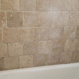 Tan, textured, square stone tiles arranged in a grid pattern installed on a bathroom wall above a white bathtub.