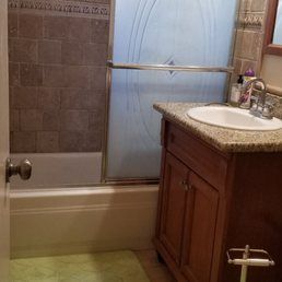 A bathroom vanity with a speckled countertop and wooden cabinet next to a bathtub with brown tiled walls.
