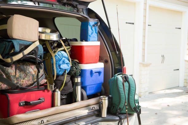 The open trunk of an SUV packed with camping gear, including coolers, bags, and a green backpack, in front of a garage.