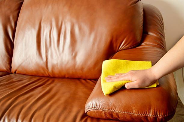 A hand wipes the arm of a brown leather sofa with a yellow cloth.