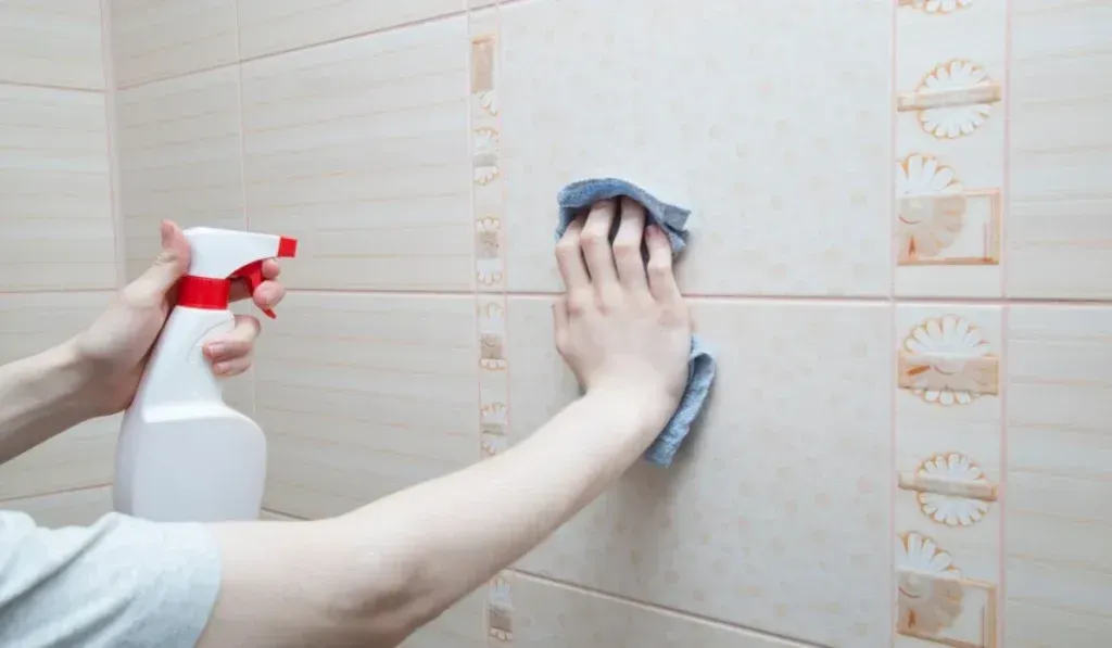 A hand holds a spray bottle while another hand wipes down light-colored tiled walls with a cleaning cloth.