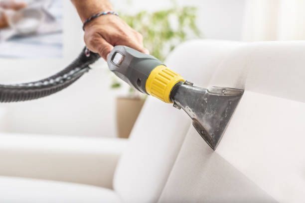 A close-up of a hand using an upholstery cleaning machine to clean a white sofa.