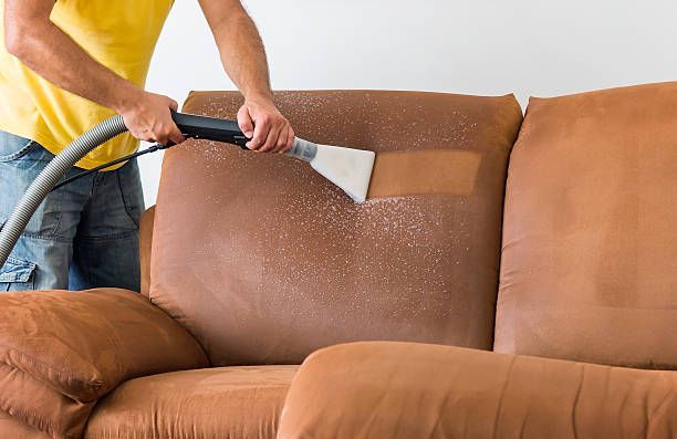 A person in a yellow shirt uses a steam cleaning tool to deep clean a brown fabric sofa.