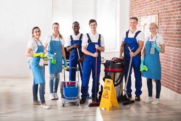 A team of cleaners in blue aprons and uniforms standing together in an office with cleaning tools and equipment.