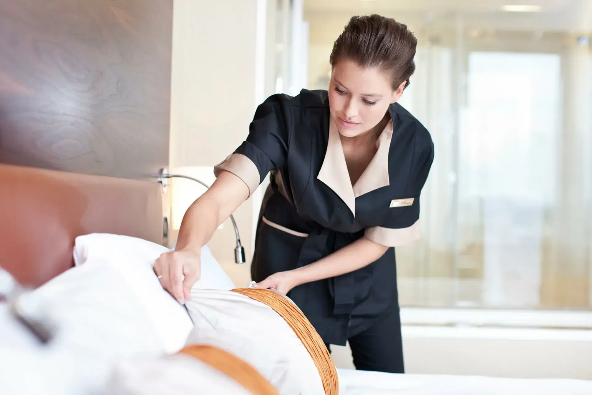 A hotel housekeeper in a black and beige uniform makes a bed, smoothing a white pillow in a bright guest room.