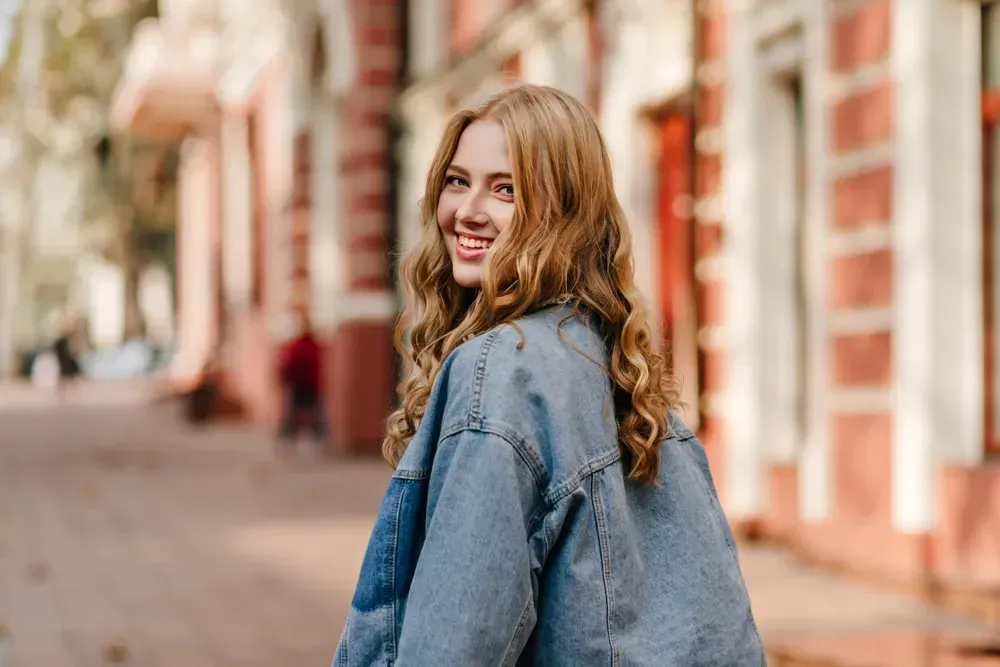 The woman is wearing a denim jacket and smiling while walking down the street.