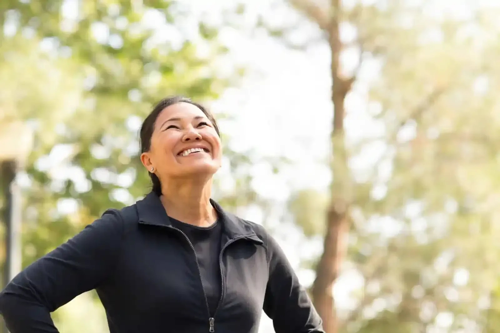 A woman is standing in a park with her hands on her hips and smiling.