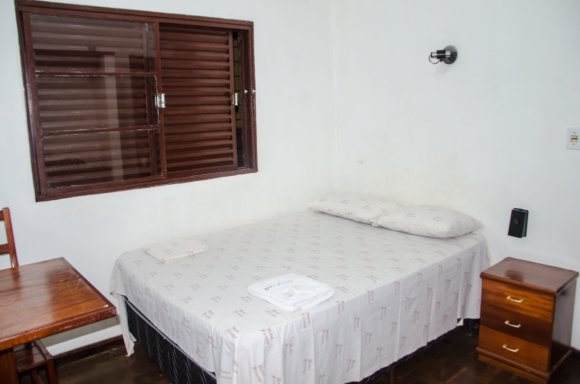 Um quarto com uma cama, mesa de cabeceira, cadeira e janela.