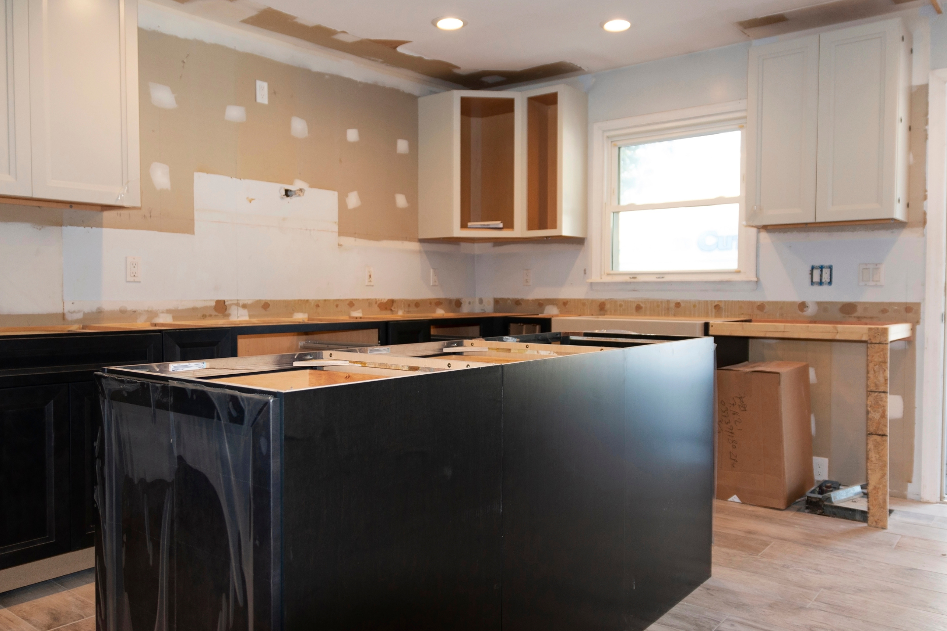 A bright kitchen under renovation with unfinished black lower cabinets and white upper cabinets. The island is partially constructed, and the walls are bare.