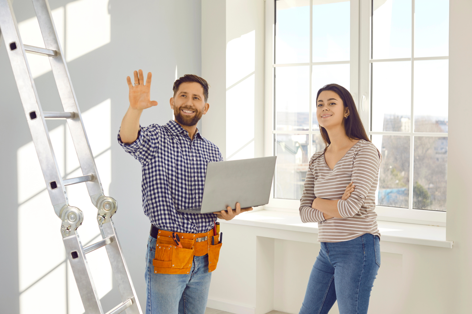 A smiling man with a laptop gestures while standing next to a woman in a room with a ladder and large windows. They appear to be discussing renovations.