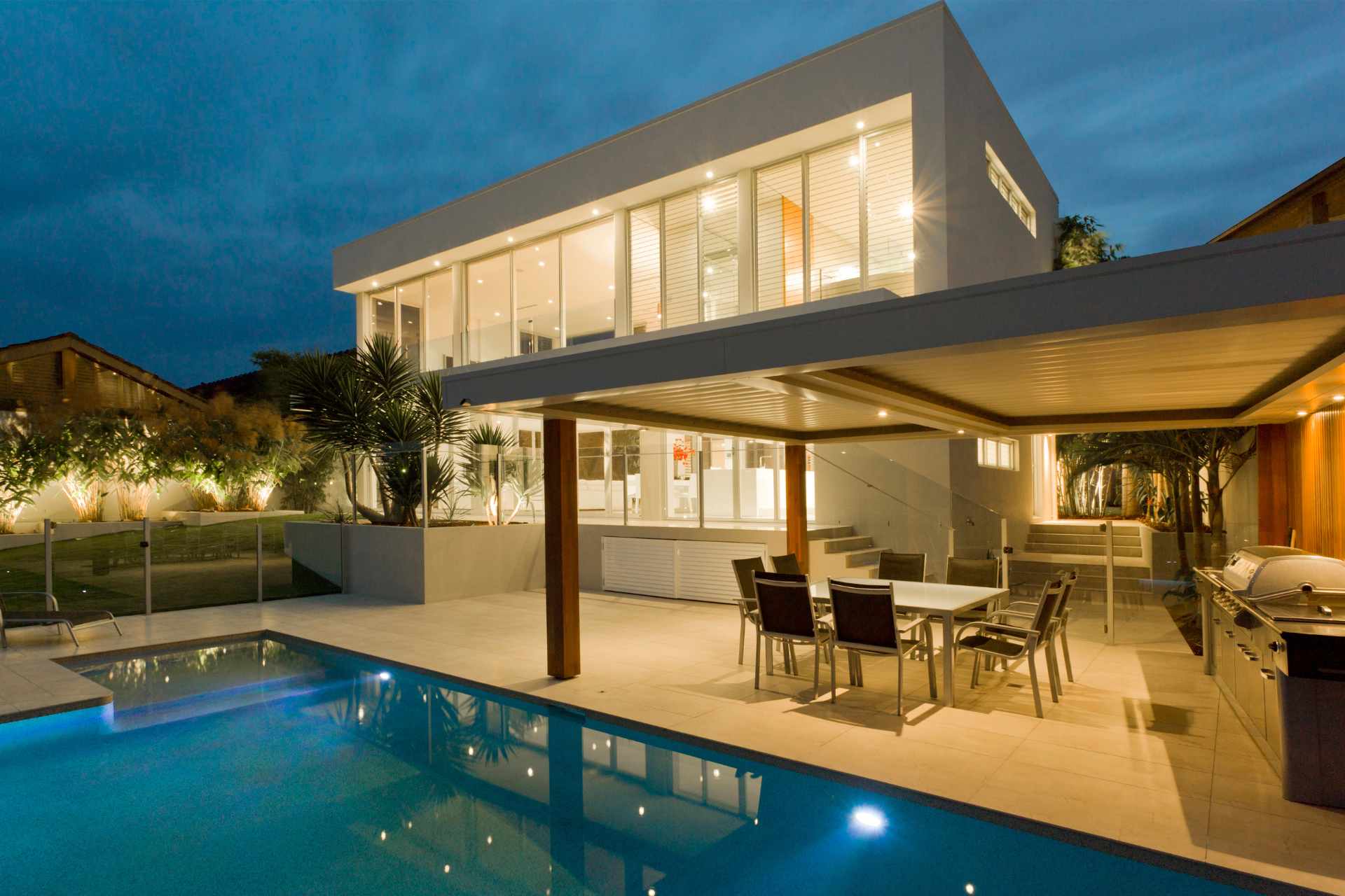Modern two-story house with large windows, glowing warmly at dusk. Features a sleek patio.