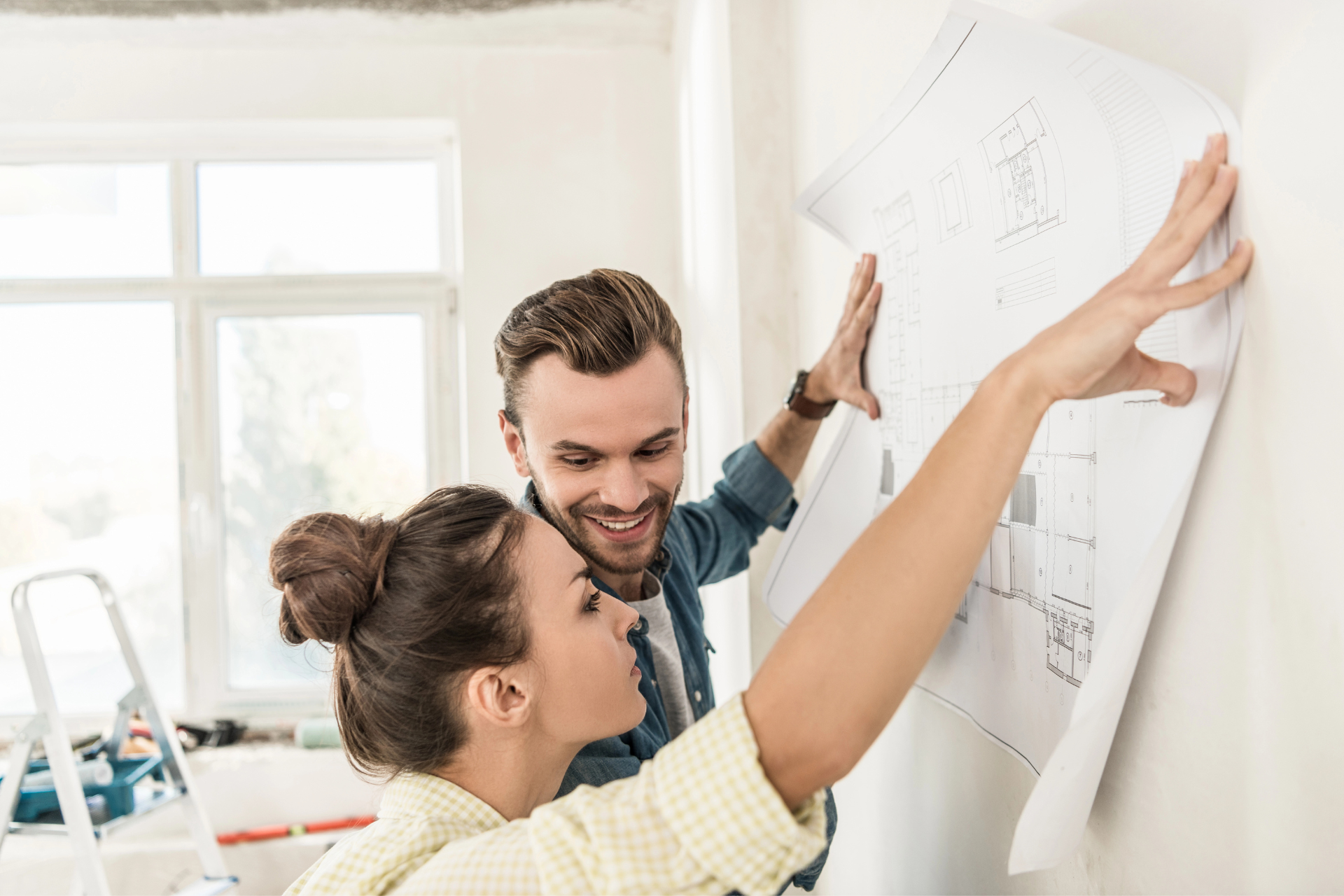 A couple examines a house blueprint pinned to a wall. The man smiles, while the woman points, looking focused. Light streams from a window, suggesting renovation.