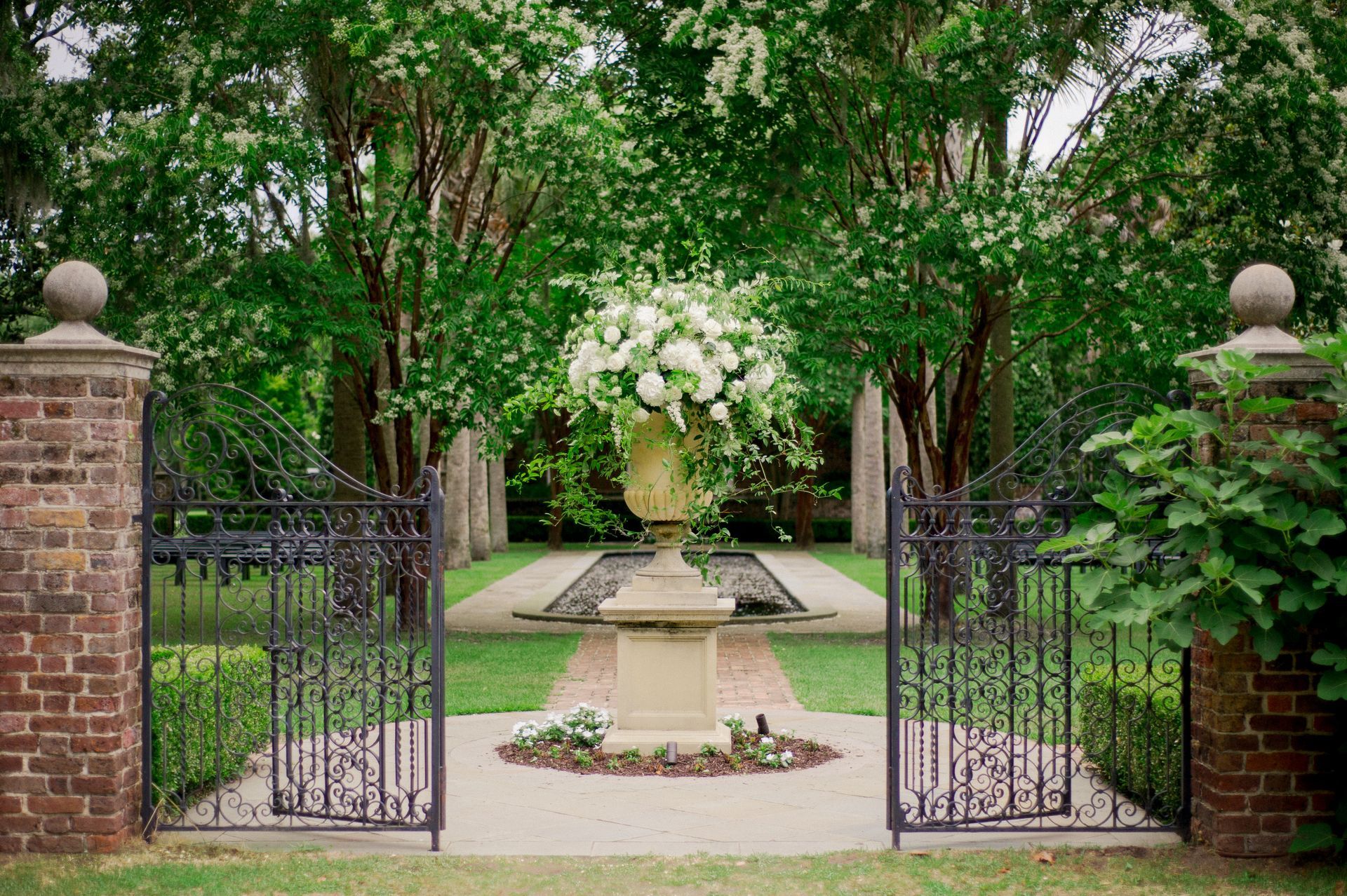 Open wrought-iron gates lead to a formal garden, featuring a large floral arrangement in a stone urn.