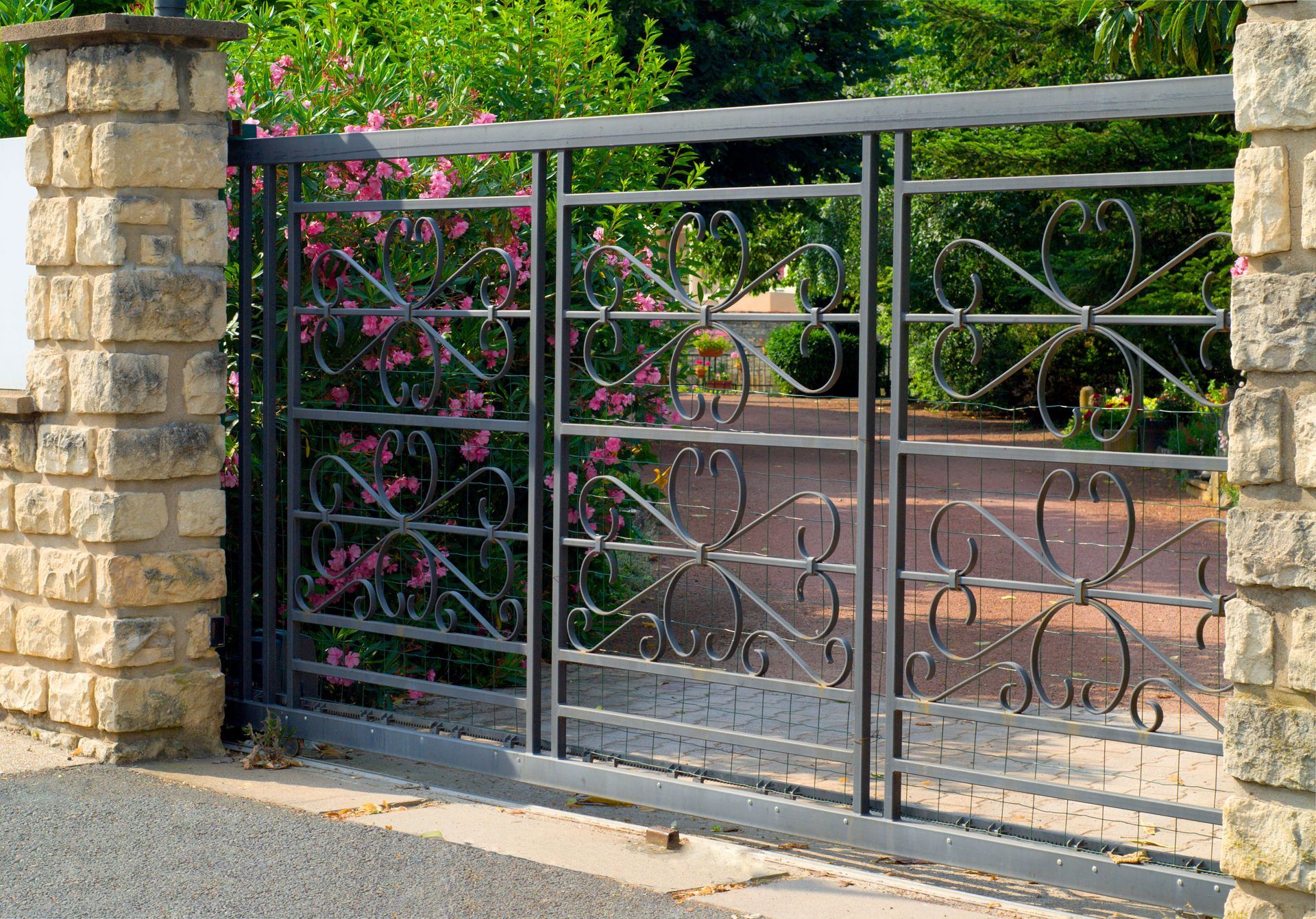 A wrought iron gate between stone pillars, set in front of a garden with pink flowers.