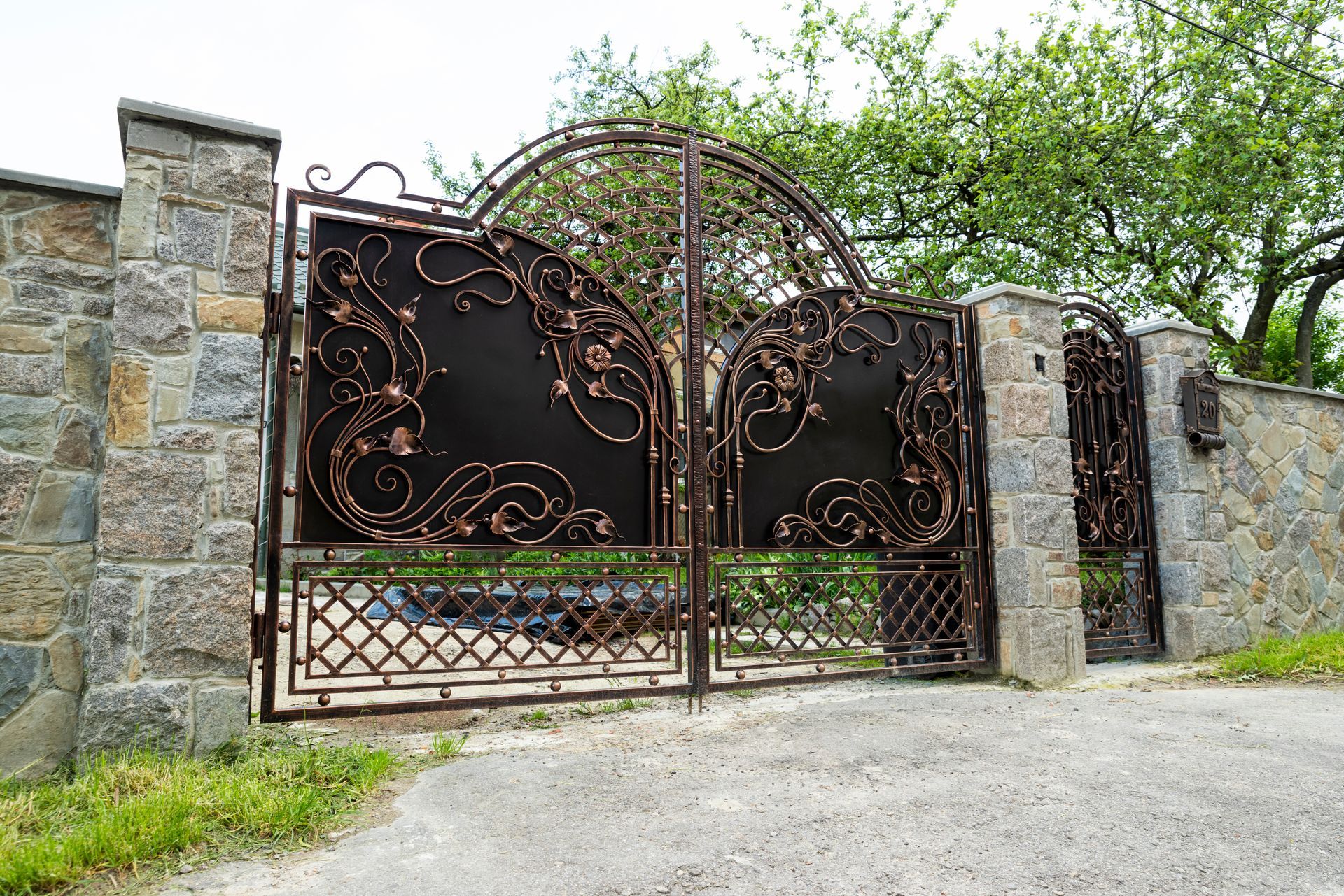 Ornate, dark metal gates in stone pillars open to reveal a driveway and greenery.