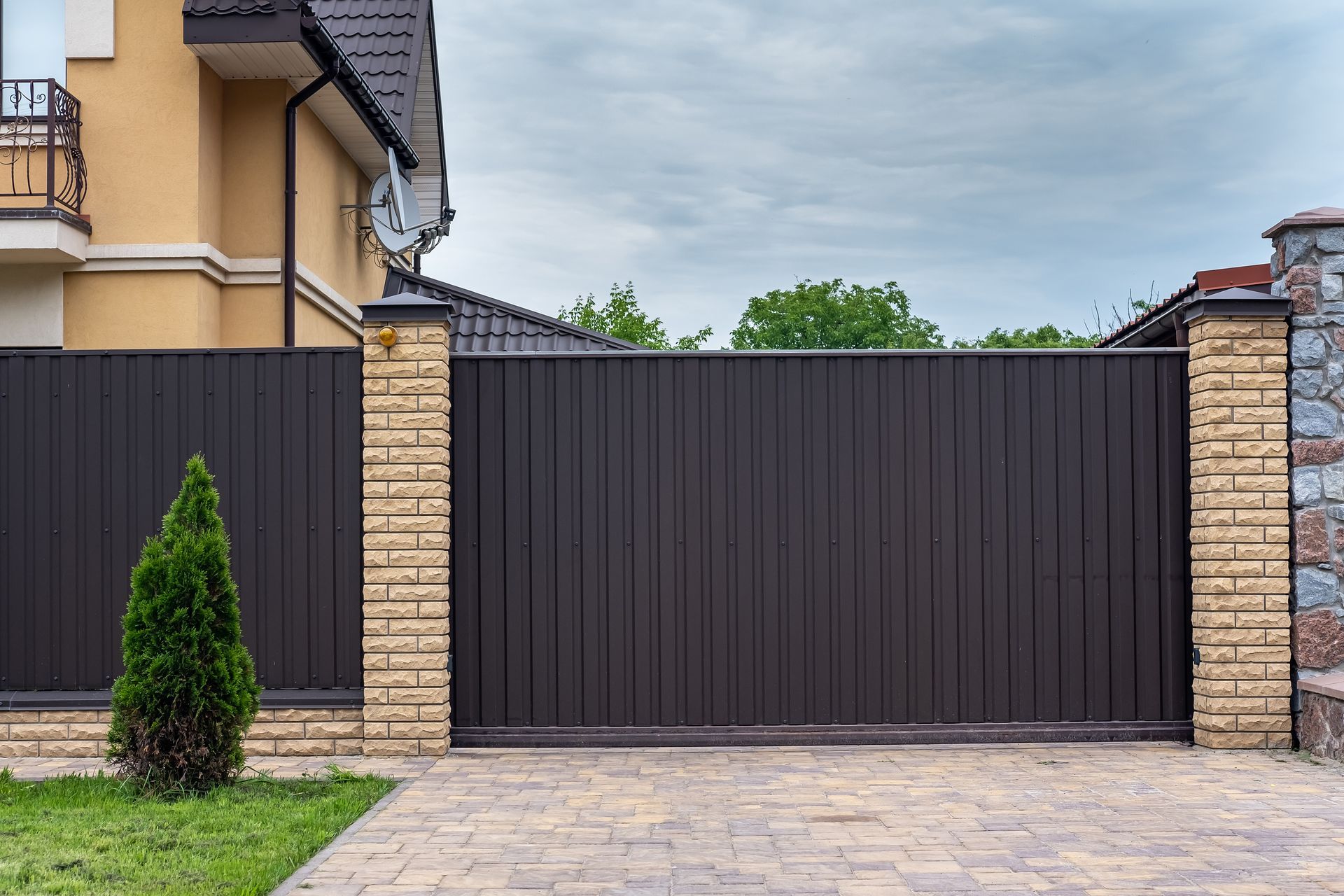 Brown metal sliding gate with brick columns, in front of a house, cloudy sky.