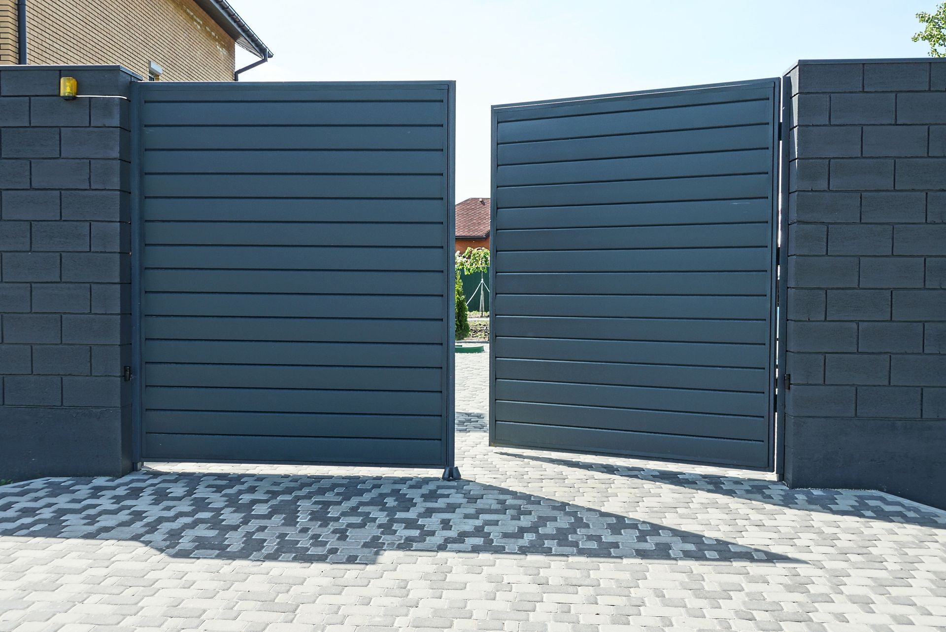 Dark gray, horizontal slatted double gate, open on gray brick pillars, on a cobblestone driveway.