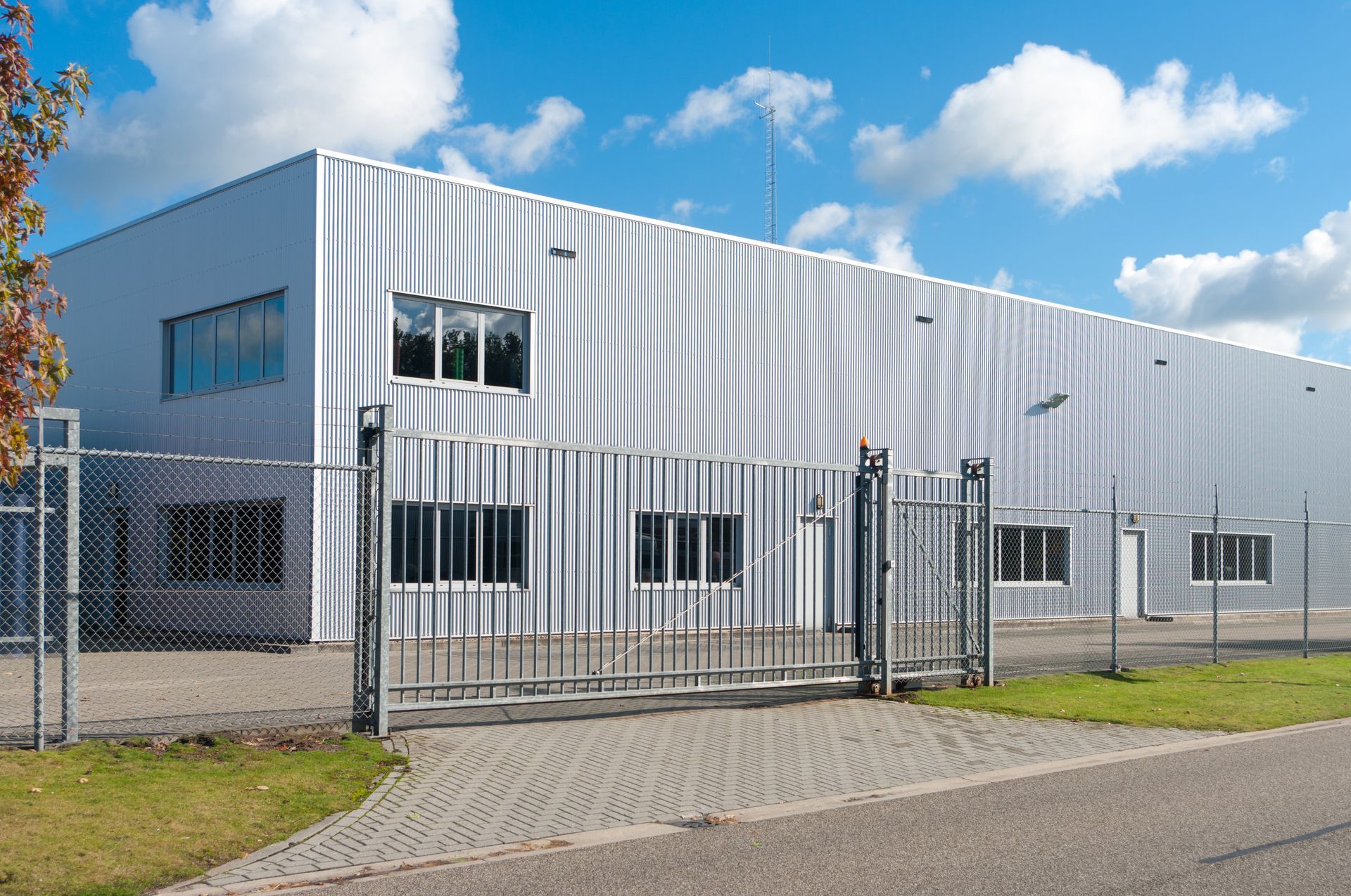 Industrial building behind a chain-link fence, with an open gate on an asphalt drive under a partly cloudy sky.