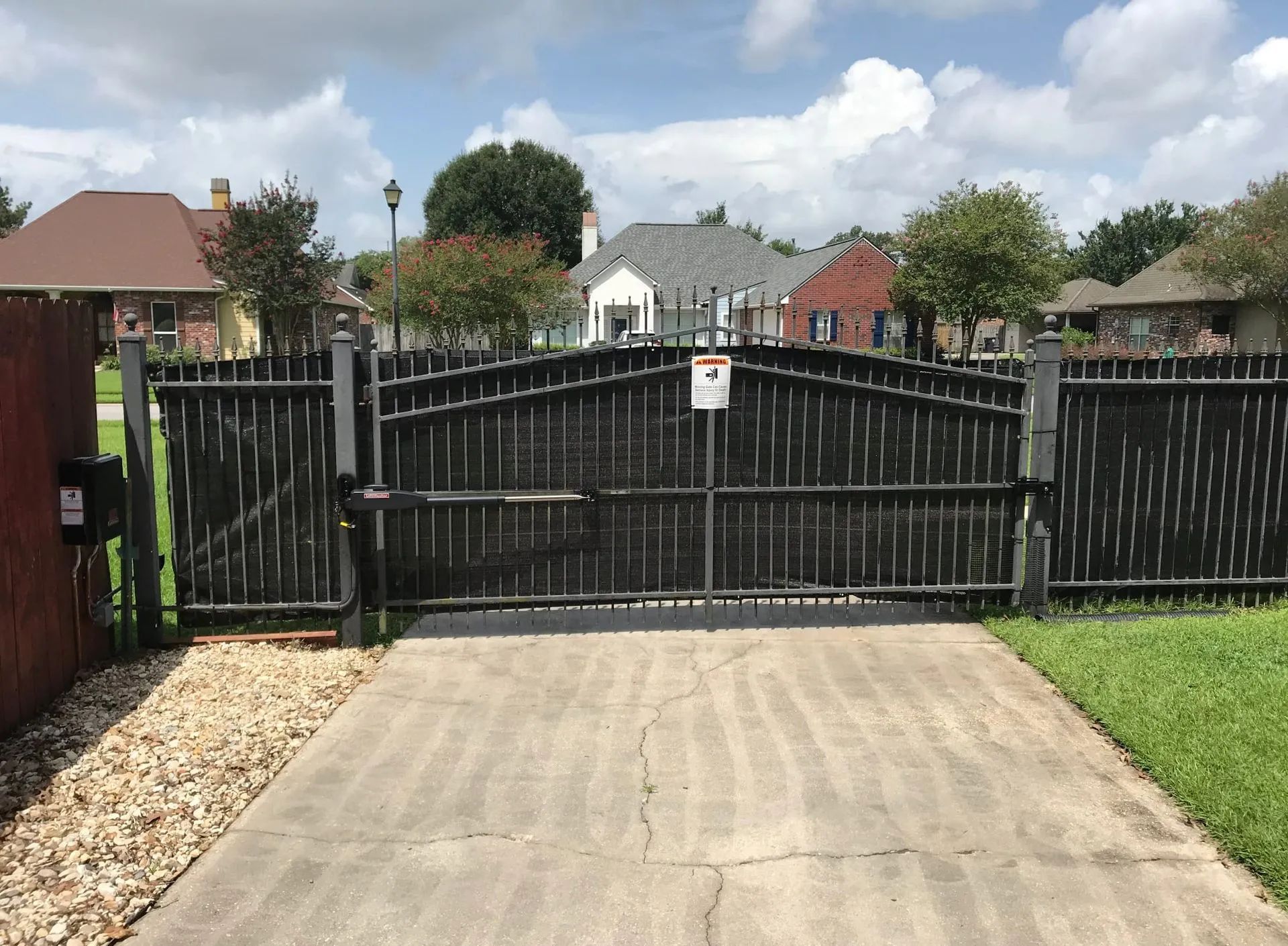 Black metal gate with security camera, blocking a concrete driveway. Houses in background.