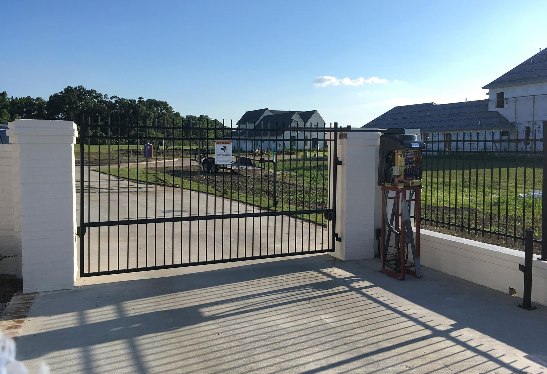 Black metal gate between white pillars, leading to a driveway, with a house in the background.