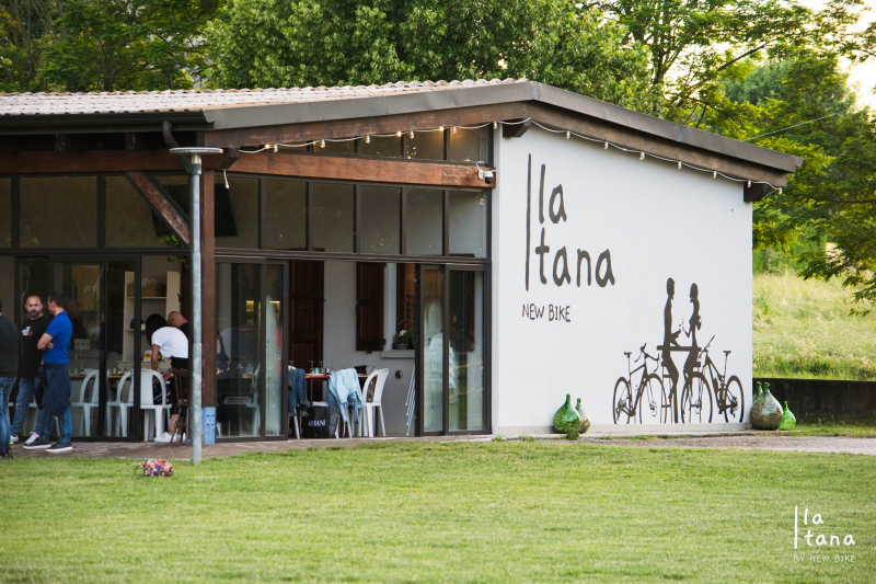 Lungo tavolo in legno apparecchiato per pranzare, con tovagliette di carta, bicchieri e posate. Vista esterna.