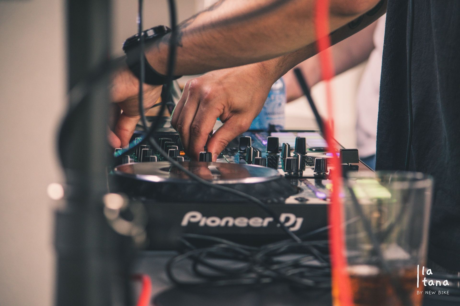 Primo piano delle mani di una persona che regola manopole e fader su un controller Pioneer DJ. Sono visibili cavi, bevande e un oggetto rosso.