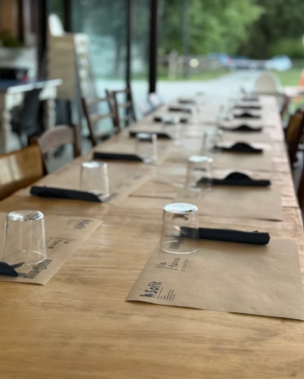 Lungo tavolo in legno apparecchiato per pranzare, con tovagliette di carta, bicchieri e posate. Vista esterna.