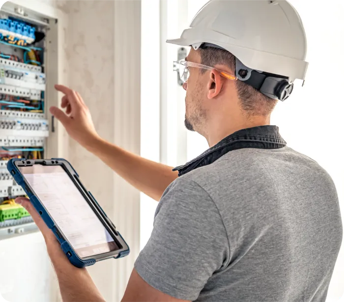 Electrician inspecting electrical panel, holding tablet, wearing hard hat and safety glasses.