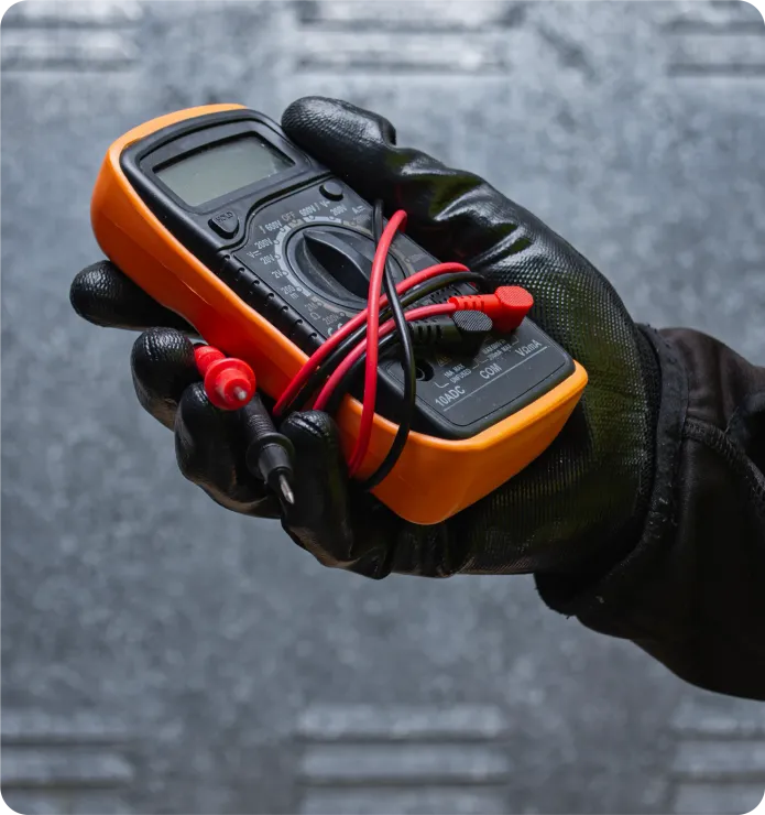 Gloved hand holding an orange and black multimeter with red and black probes, in front of a gray surface.