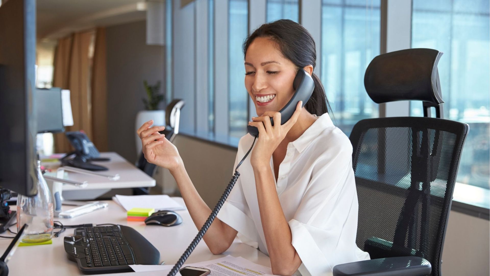 Woman in white shirt talking on phone at a desk in an office setting.